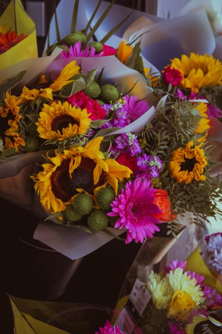 A vibrant floral bouquet featuring bright yellow sunflowers with large dark centers, pink gerbera daisies, and small purple filler flowers, complemented by green billy balls and grayish-green foliage.