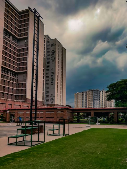 The image shows a modern urban park with a well-maintained grassy area and a paved seating zone featuring black metal benches with wooden slats. In the background, there are tall residential or office