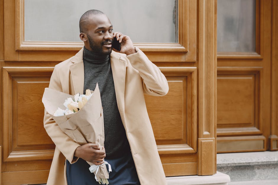 A man with a neatly trimmed beard and short hair, dressed in a beige overcoat and dark turtleneck, holding a bouquet of fresh white flowers wrapped in brown craft paper, standing outdoors in front of