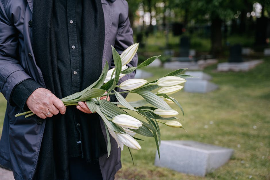 A person wearing a dark coat and black shirt is holding a bouquet of white lilies with long green leaves, arranged with some flowers facing outward and others tucked inward. The bouquet is wrapped in 