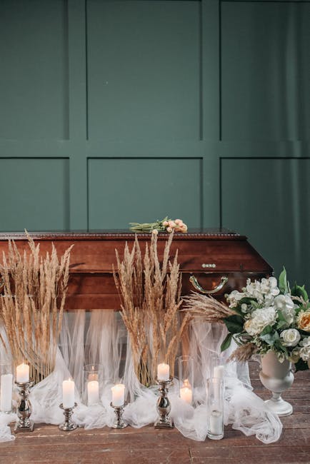 A floral tribute scene featuring white roses, peonies, and greenery arranged in a large, elegant bouquet placed on a white vase. The bouquet is positioned on a table with a green paneled wall backgrou