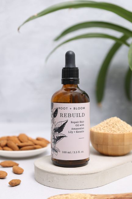 A close-up of a small amber glass bottle of 'Rebuild' repair hair oil with a black dropper cap, positioned on a white, oval-shaped stone slab. In the background, there is a green leafy plant with long