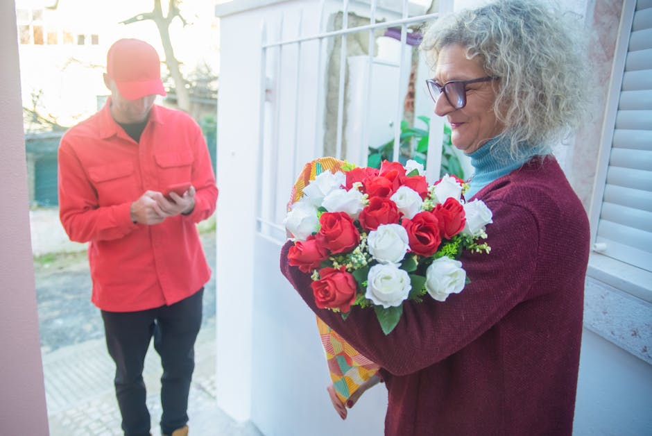 A woman with curly gray hair, glasses, and a light blue turtleneck sweater under a burgundy cardigan, is holding a vibrant bouquet of fresh roses in red and white, wrapped in decorative paper. She sta
