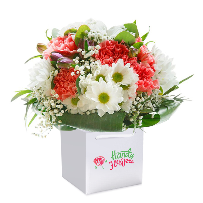 Presented in a pristine, square white gift bag, this radiant bouquet gleams as the embodiment of grace and celebration in Chase Cross. The main attraction is the collection of soft coral-pink and peach carnations, their dense, ruffled petals forming plush, billowy shapes that nearly spill over the bag's edges. Bright white daisies, with eye-catching lime-green centers and crisp petals, sit among the carnations, catching the light and reflecting a spirit of carefree happiness reminiscent of lazy afternoons in neighbouring Havering Country Park. Sprigs of baby's breath weave a fine, gossamer texture throughout, adding weightlessness and charm. Accents of unopened, purple-tipped buds nestle to the left-possibly heralding alstroemeria's delicate arrival-infusing the bouquet with quiet anticipation. Framing it all is an abundant collar of lush, deep green foliage whose smooth leaves cup the arrangement in a protective embrace, each leaf shining gently as if just misted. The sturdy ribbon handles arch gracefully above, ready to carry good wishes to a friend or loved one for any celebratory purpose: birthdays, anniversaries, or tokens of gratitude. The pure white backdrop isolates each colour and shape for maximum effect, allowing the vibrancy of the arrangement to speak for itself-reflecting the skill and expertise of Chase Cross's dedicated florists and their commitment to delivery of only the freshest blooms to the wider community.