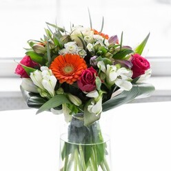 This radiant floral arrangement, expertly designed in Chase Cross, exudes both warmth and sophistication-an ideal centrepiece for gatherings in the heart of our community. The bouquet commands attention with its solid core of plush gerbera daisies-two ablaze with vivid orange, two washed in creamy-peach, each delicate petal circling a rich espresso-brown centre. Velvet-furled magenta roses cluster nearby, lending an elegant splash of deep colour reminiscent of twilight skies above Mawney Brook. Ivory alstroemeria blooms, punctuated by dainty white fillers, lend an ethereal touch and suggest the effortless beauty of untended wildflowers bordering Chase Cross's leafy avenues. Sweeping arcs of broad, waxy aspidistra leaves enfold the bouquet's base, their lustrous green providing visual strength and unity, while slender, pointed leaves and airy sprigs peak between blossoms, enhancing the sense of volume and organic texture. Housed in a tall, slender glass vase-its transparency revealing a tangle of vibrant green stems-the composition feels at once tailored and wild, channeling the contrasting energy of nature and human craft. With glowing morning light gently enhancing the blooms, the bouquet radiates an inviting fragrance, blending sweet rose and gentle lily notes. A perfect gift to mark a milestone or express appreciation, this floral creation reflects both the artistry and heartfelt touch of Chase Cross Flowers, unwavering in commitment to freshness and local charm.